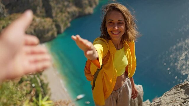 POV happy woman hiker in yellow shirt gives hand to her teammate on edge of cliff on mountain top, Butterfly Valley in Oludeniz, Fethiye western Turkey. view on turquoise sea from above