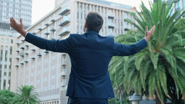 man spread hands in the air praising God standing outdoors. businessman spreading his hands apart to welcome. businessman joyfully spreads hands stands against the backdrop of skyscrapers back view