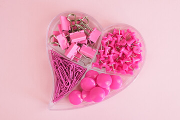 Stationery in pink color in a transparent plastic box in the shape of a heart on a pink background. Paper clips, push pins, binder clips in pink design. Back to school concept. Top view