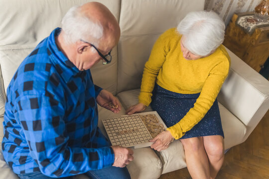 Senior European married couple playing a board game scrabble while sitting on a couch in the living room together. High quality photo