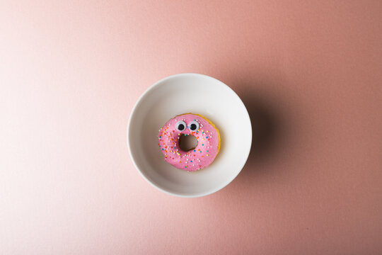 Directly above view of pink donut with googly eyes in plate around copy space on colored background
