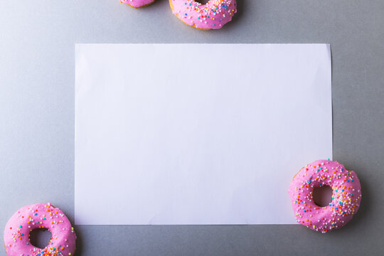 Directly above view of blank white paper with copy space amidst pink donuts on gray background