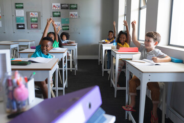Obraz na płótnie Canvas Smiling multiracial elementary school students with hands raised sitting at desk in classroom