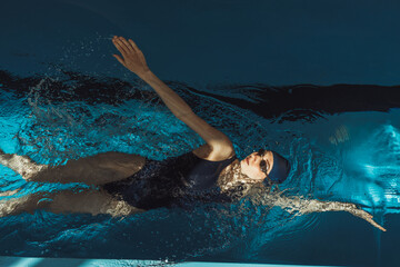From above of professional female swimmer in swimsuit goggles and cap doing backstroke while...