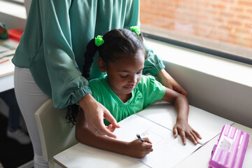 Midsection of caucasian young female teacher assisting african american schoolgirl studying at desk