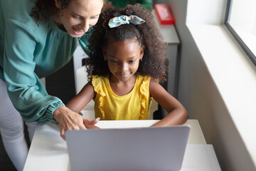 Smiling caucasian young female teacher teaching computer to african american elementary girl at desk