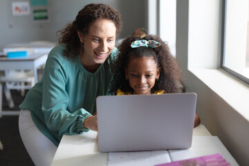 Smiling caucasian young female teacher teaching laptop to african american elementary schoolgirl
