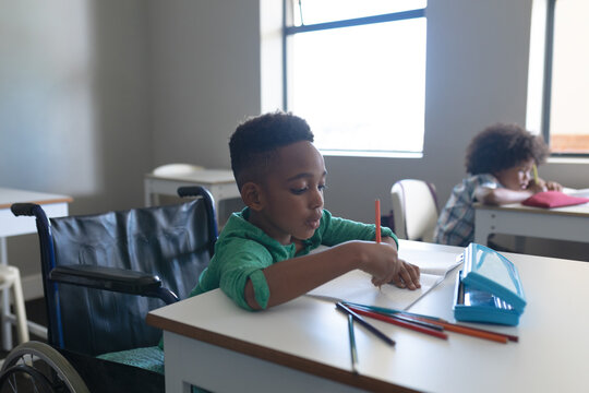 African american elementary boy writing on book while sitting on wheelchair at desk in classroom