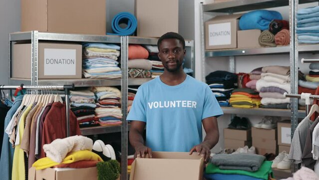 Portrait of male volunteer standing at the warehouse and looking at the camera while sorting and iterating clothes for donations. Belongings at the shelves at the background. Humanitarian aid concept