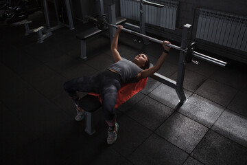 Top view shot of a fitness woman doing barbell bench press in dramatic gym lighting