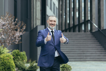 Successful senior gray haired businessman boss, outside office building smiling and looking at camera, experienced investor showing middle finger up, man in business suit