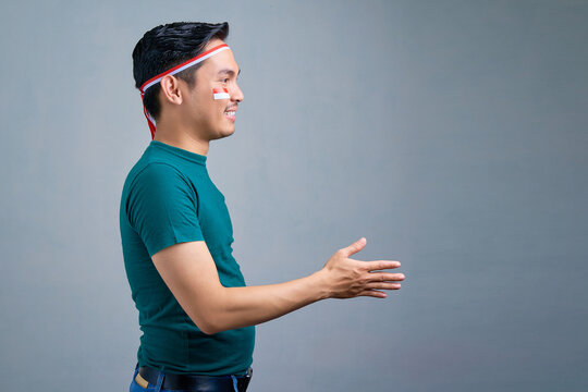 Smiling young Asian man in casual t-shirt stretching hand for handshake isolated on grey background. indonesia independence day celebration concept - Powered by Adobe