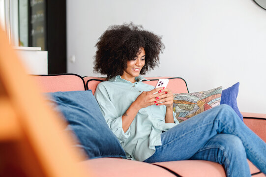 Charming multiethnic woman with Afro hairstyle using smartphone at home, multiracial girl sitting on couch, spending time online, texting messages, scrolling news feed, sharing post in social networks