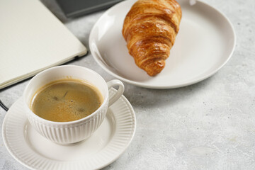 A croissant and a cup of coffee on white plates on grey working desk in the office with laptop and notebook, copy space for text