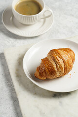French soft croissant on a white plate on a marble tray, a cup of black coffee in white sophisticated cup
