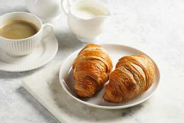 Two french soft croissants on a white plate on a marble tray, a cup of black coffee in white sophisticated cup, milk, sugar, jam