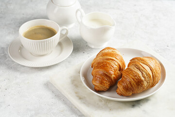 Two french soft croissants on a white plate on a marble tray, a cup of black coffee in white sophisticated cup, milk, sugar, jam