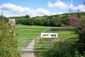 A hand painted shut gate sign attached to a metal gate at the entrance to a footpath running through green countryside in summer