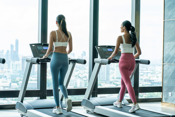 Fototapeta na wymiar Two beautiful Asian friends exercise together in sportswear, walk and run on the treadmill in the rooftop gym.