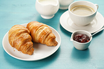 Two french soft croissants on a white plate on a blue wooden table, a cup of black coffee in white sophisticated cup, milk, sugar, jam