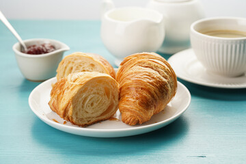 Two french soft croissants on a white plate on a blue wooden table, a cup of black coffee in white sophisticated cup, milk, sugar, jam