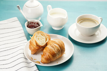 Two french soft croissants on a white plate on a blue wooden table, a cup of black coffee in white...