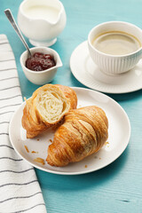 Two french soft croissants on a white plate on a blue wooden table, a cup of black coffee in white sophisticated cup, milk, sugar, jam
