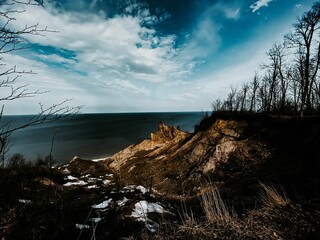 Scenery of a rocky cliff with leafless trees and melting snow under a blue sky and white clouds