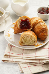 Two french soft croissants with jam on a white plate on a checkered napkin, a cup of black coffee in white sophisticated cup, milk, sugar