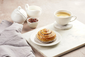 A round cinnamon pastry on a white plate on marble board, cup of coffee, jam, milk