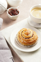 A round cinnamon pastry on a white plate on marble board on light pink background, cup of coffee, jam, milk