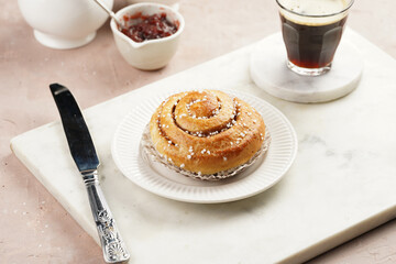 A round cinnamon pastry on a white plate on marble board on light pink background, cup of coffee, jam, milk