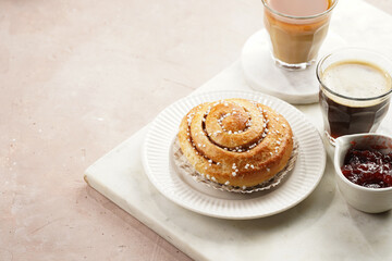 A round cinnamon pastry on a white plate on marble board on light pink background, cup of coffee, jam, milk