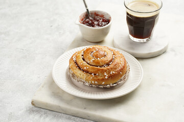 A round cinnamon pastry on a white plate on marble board on light pink grey, cup of coffee, jam, milk