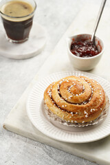 A round cinnamon pastry on a white plate on marble board on light pink grey, cup of coffee, jam, milk