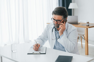 Man giving information by phone. Professional medical worker in white coat is in the office