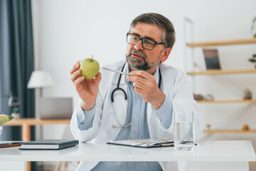Naklejka na ściany i meble Male doctor is with an apple. Professional medical worker in white coat is in the office