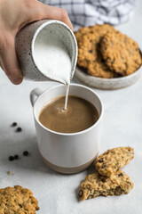 Close-up of a hand pouring foamed milk into espresso coffee in a coffee cup. On a light background with a scattering of chocolate beans and oatmeal cookies. The concept of a homemade breakfast.