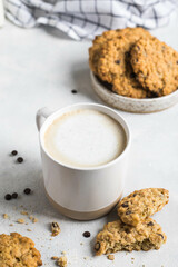 Close-up of a cup of coffee with milk foam. On a light background with a scattering of chocolate beans and oatmeal cookies. The concept of a homemade breakfast.
