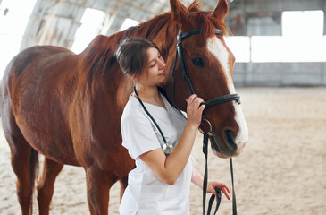 Brown colored hair. Female doctor in white coat is with horse on a stable