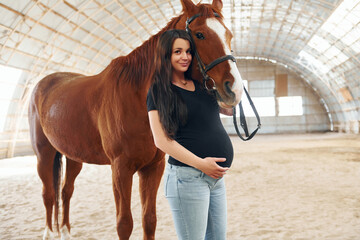 Looking at the camera. Beautiful pregnant woman in casual clothes is with the horse on a stable