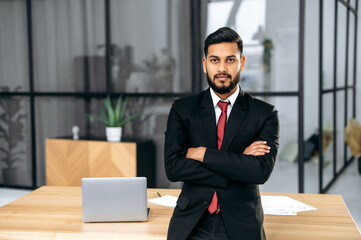 Portrait of elegant serious confident successful Indian or Arabian businessman in a suit, lawyer, male entrepreneur, stands in a modern office near the desktop with crossed arms, looks at the camera