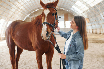 Smiling and enjoying. Young woman in jeans clothes is with horse on a stable