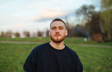 Positive young man with a beard in dark clothes stands in the park at sunset in the evening and looks at the camera.