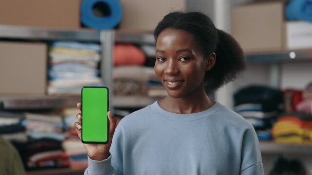 Portrait view of the multiracial woman holding smartphone with green mock up screen and looking to the camera while standing at the reuce center. Second hand concept