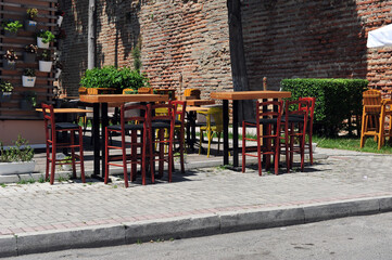 Empty tables and chairs in Albanian sidewalk cafè.  Flower arrangements in buckets on the wall. Brick wall in the background.