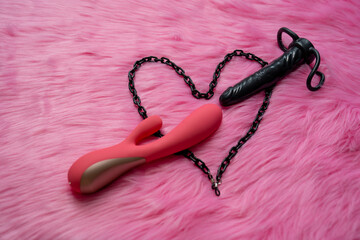 black dildo and pink vibrator sex toys next to black chain heart, on the pink fur background of bedroom. Concept of heart and arrow romance