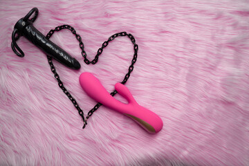 black dildo and pink vibrator sex toys next to black chain heart, on the pink fur background of bedroom. Concept of heart and arrow romance