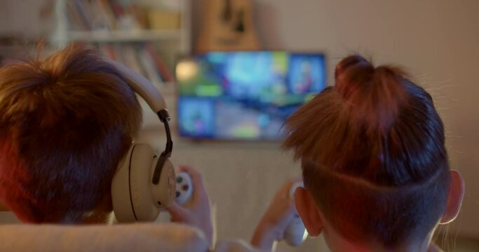 The backs of the heads of children in front of the TV. They are holding video game controllers in their hands. On screen in bokeh action game. Spending free time on holidays.