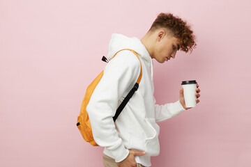 a nice man stands on a pink background in a white hoodie with a backpack on his back, holding a glass with a drink, sideways to the camera looking into the glass